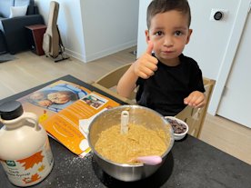 A photography of a young boy sitting next to a bowl of muffin mix smiling and giving a thumbs up
