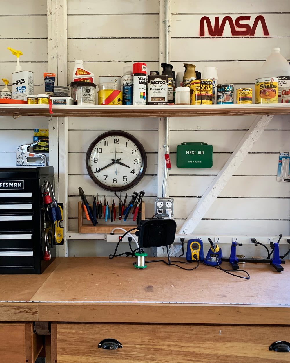 a wooden desk setup with an assortment of tools and finishes, a clock, a first aid kid, and a painted NASA logo