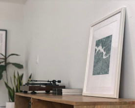 artwork in a white matte frame on a tabletop with books, a record player, and plants visible