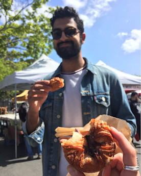 a hand holds a kouign-amann pastry with Neil eating another kouign-amann in the background