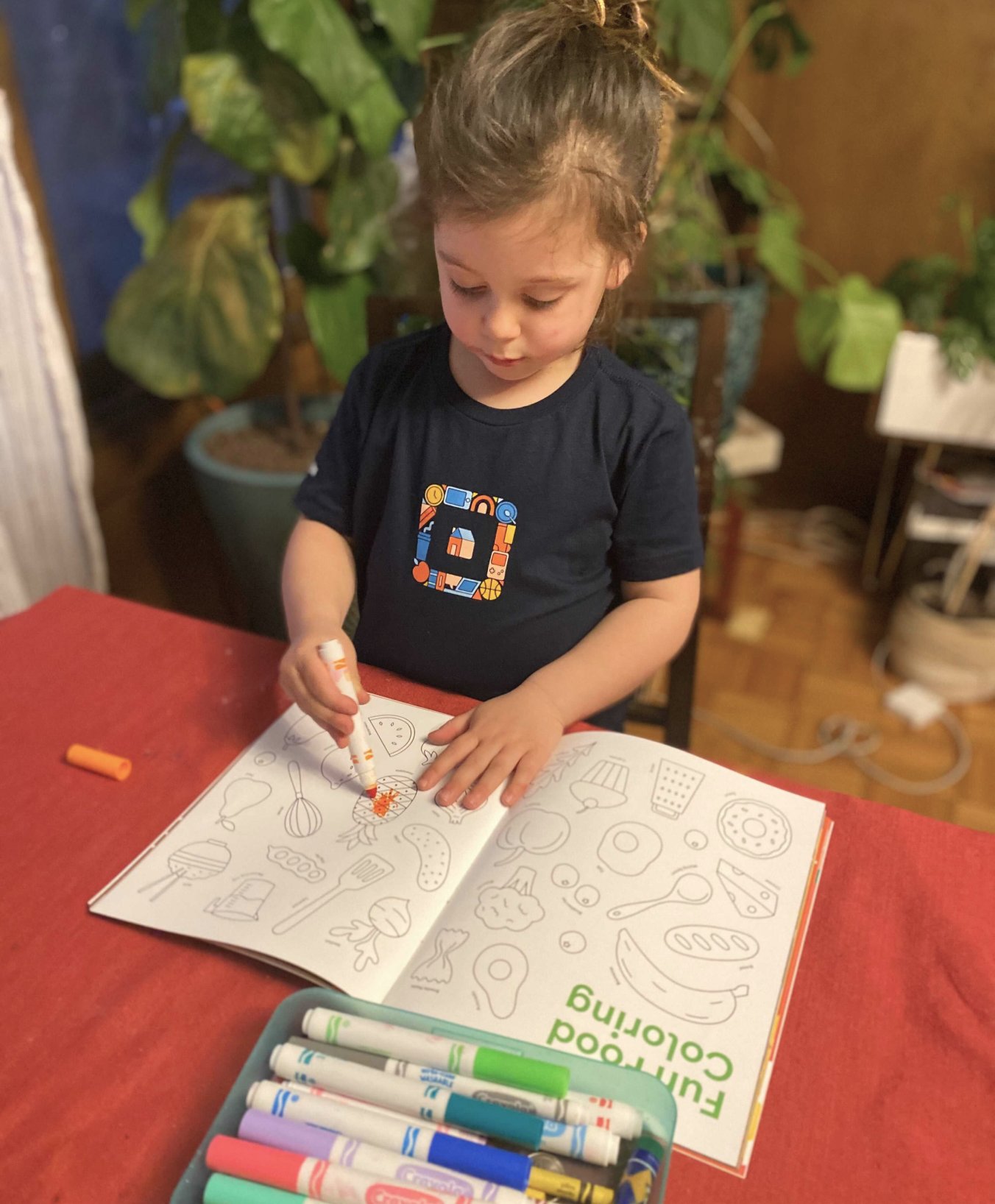 A young girl wearing a square t-shirt coloring a page of the cookbook with an orange marker
