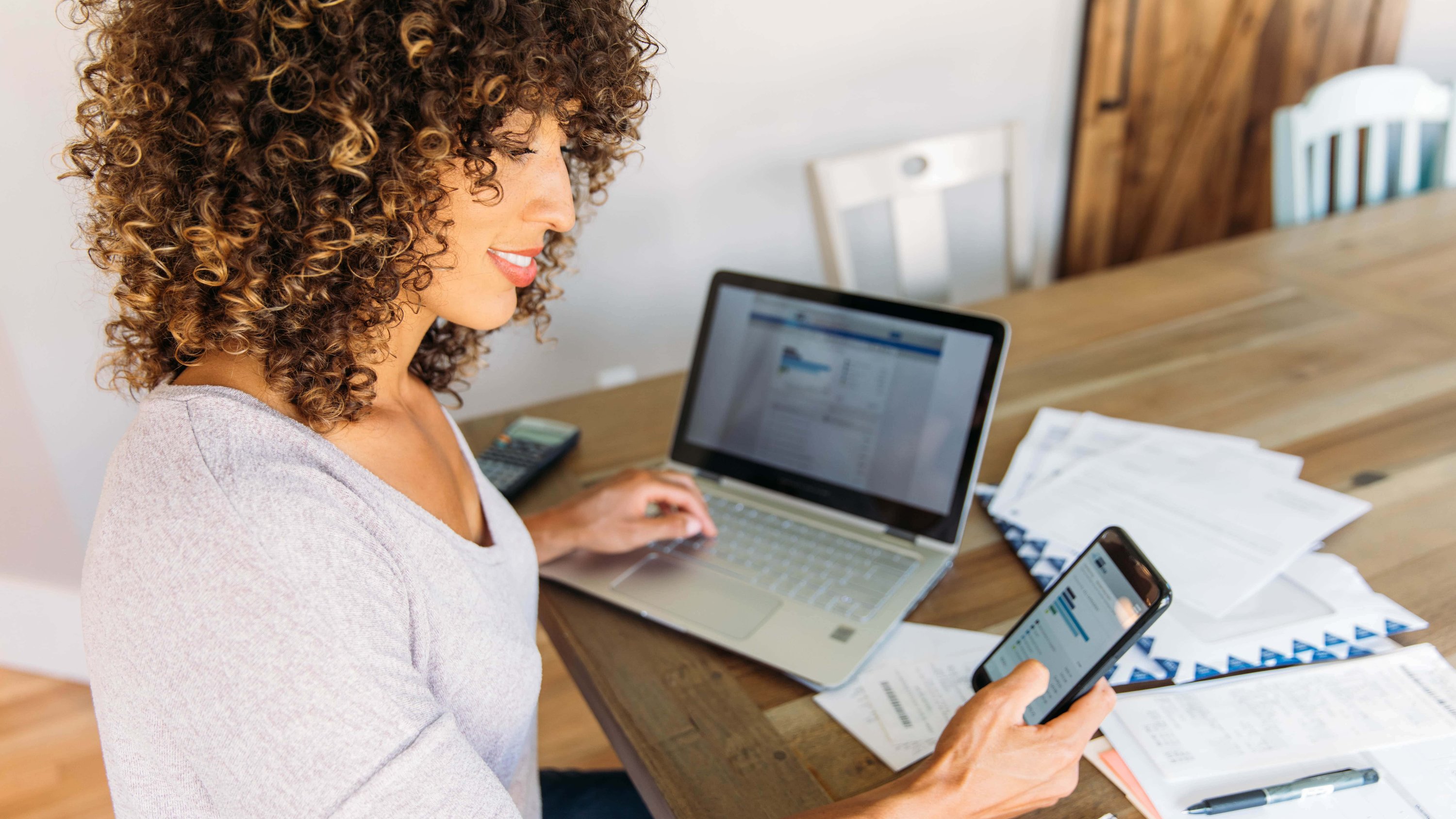 Surrounded by paper work, a business owner uses their laptop, smartphone, and calculator to manage their business transactions.