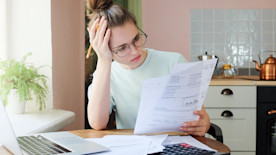 A woman working from her kitchen table reads over her bank statements with her laptop and calculator nearby.