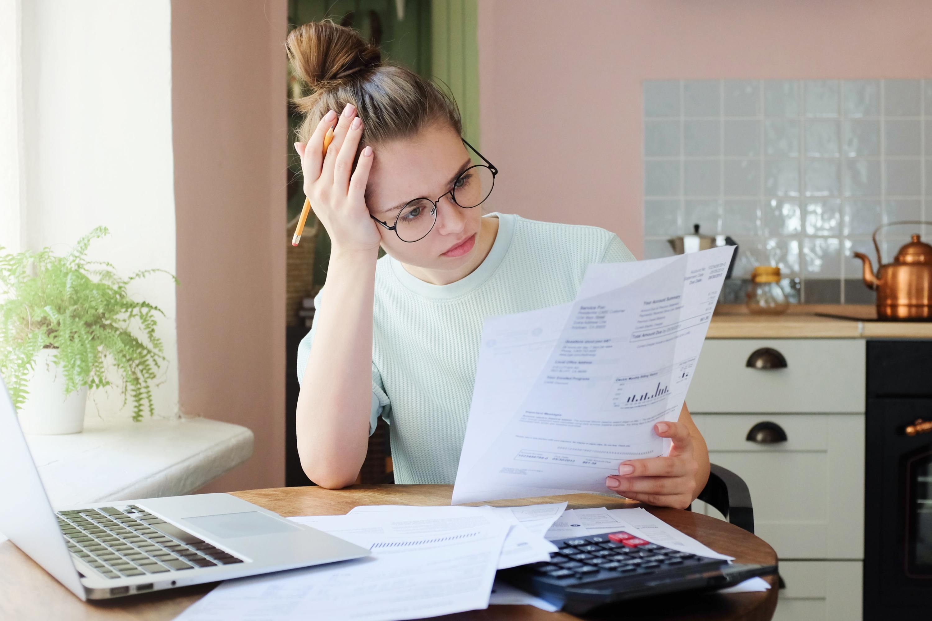 A woman working from her kitchen table reads over her bank statements with her laptop and calculator nearby.