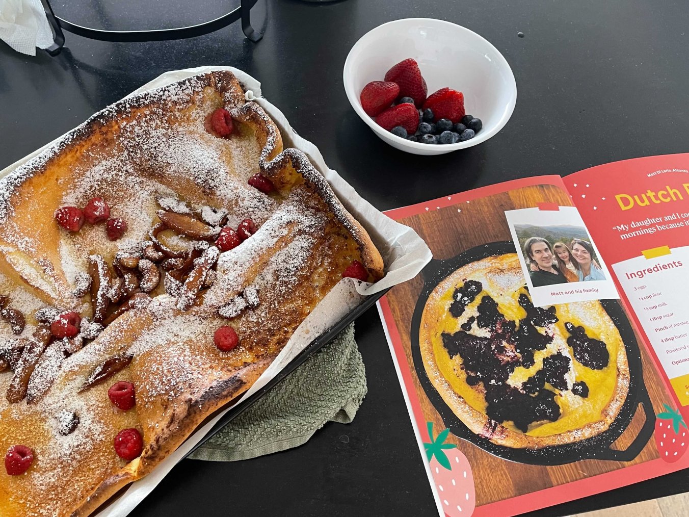 An aerial view of a dutch pancake with raspberries and powdered sugar next to the dutch pancake spread in the cookbook all on a black countertop