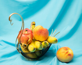 a still life photo of fruits in a decorative birdlike brass basket on a blue fabric spread