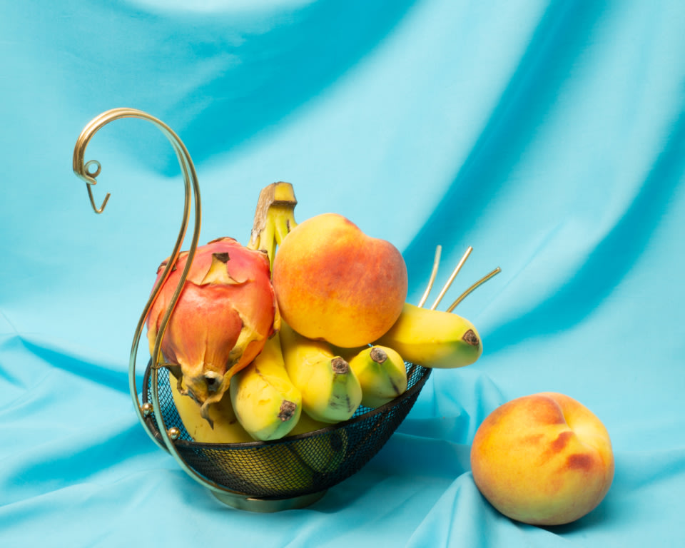 a still life photo of fruits in a decorative birdlike brass basket on a blue fabric spread