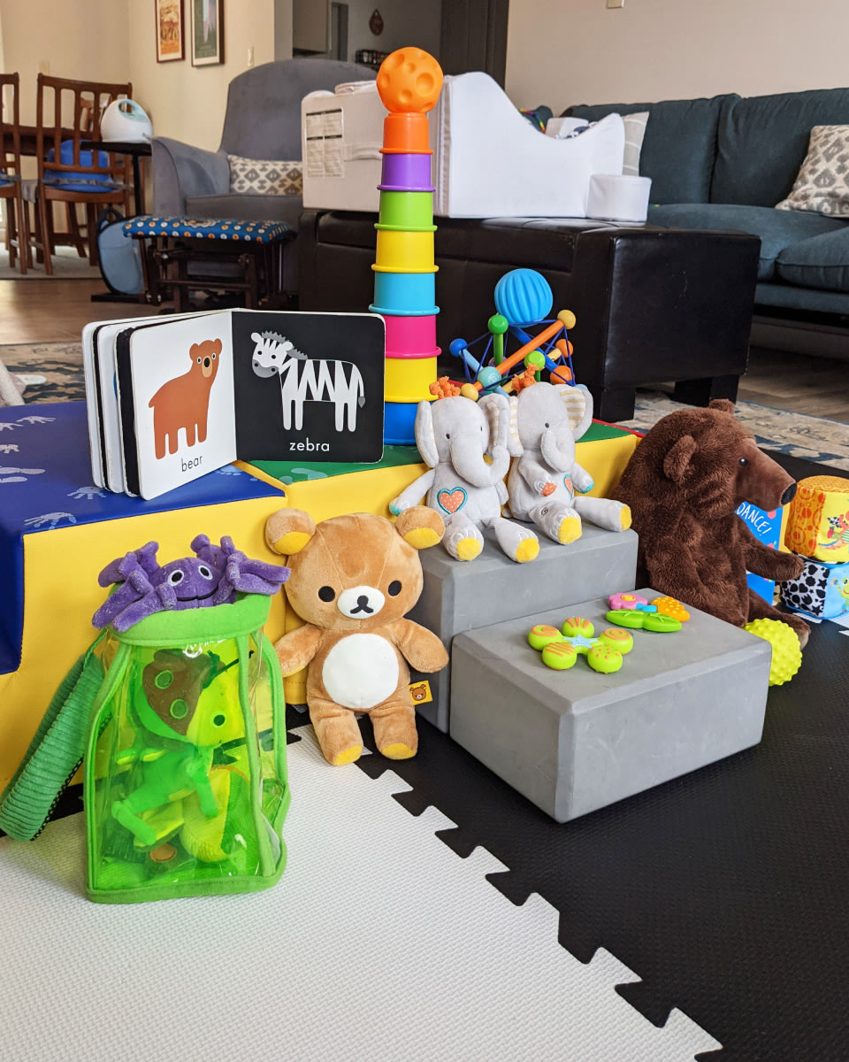 a stack of toys, blocks, stuffed animals, and a book on a puzzle mat in a living room