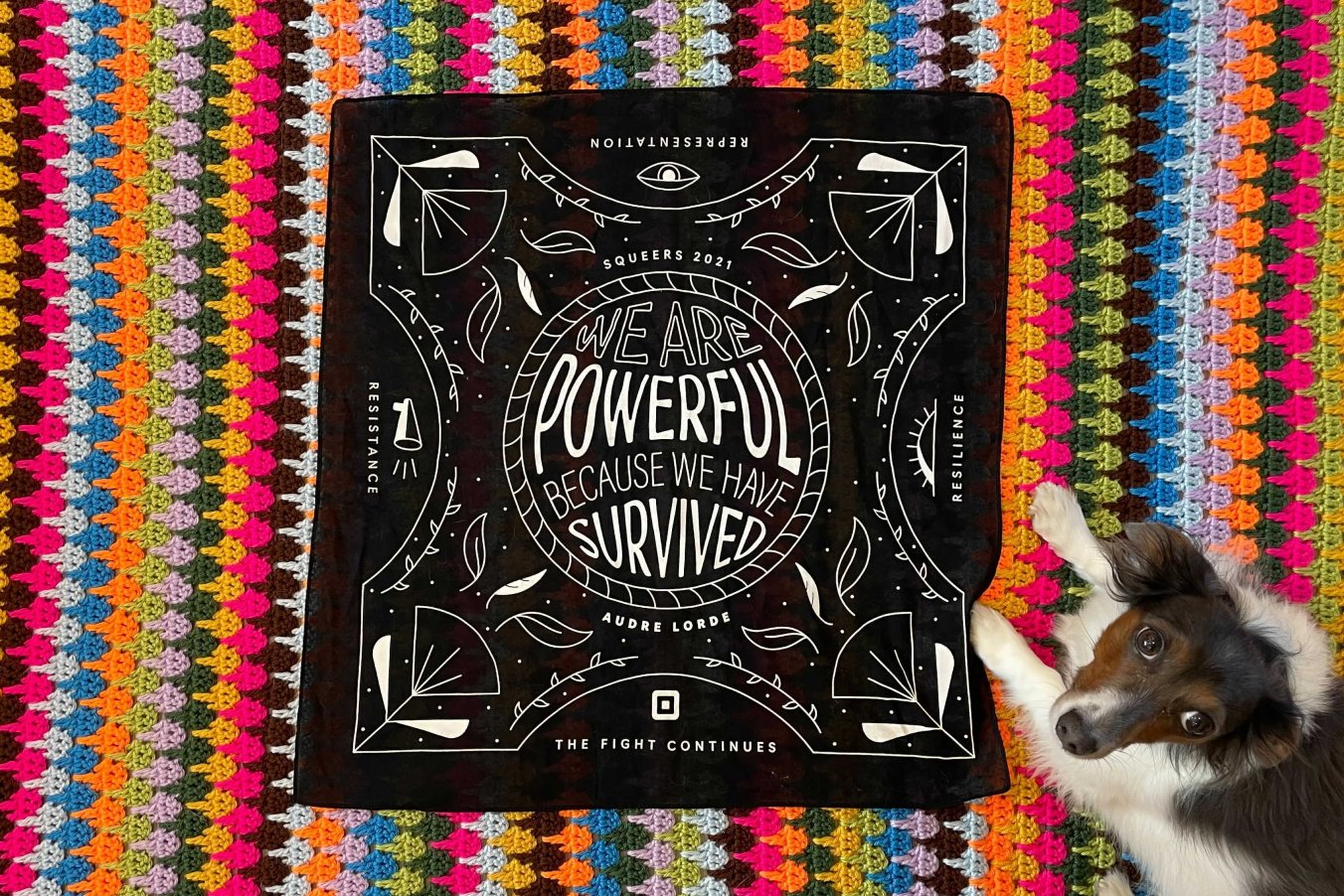 The bandana laid flat on a colorful knitted quilt with a small dog laying on top of both
