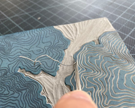 an obscured hand uses a metal tool to extrude a pattern from a wood block