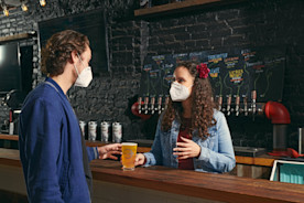 A masked patron at a bar ordering a beer from a masked bartender