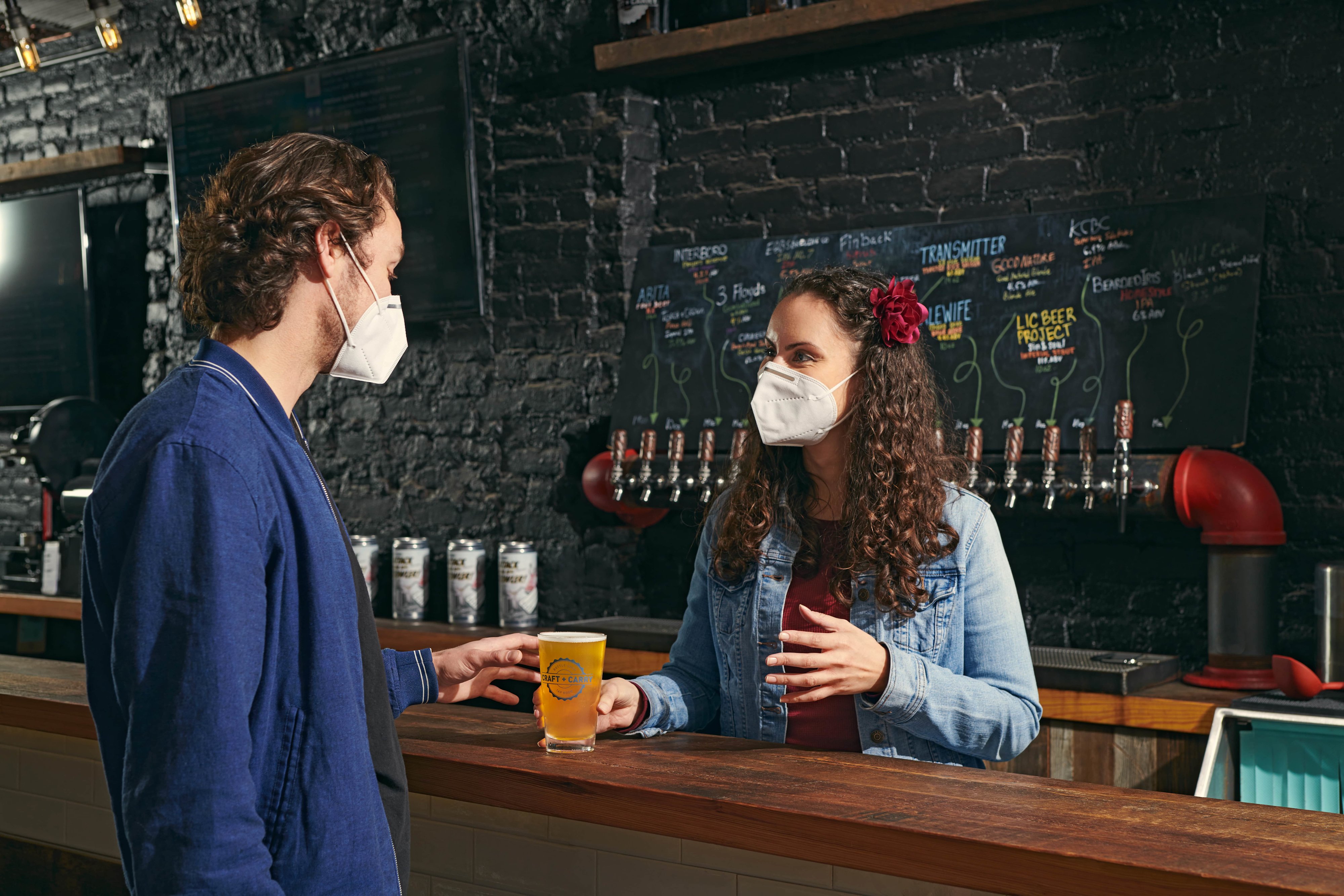 A masked patron at a bar ordering a beer from a masked bartender
