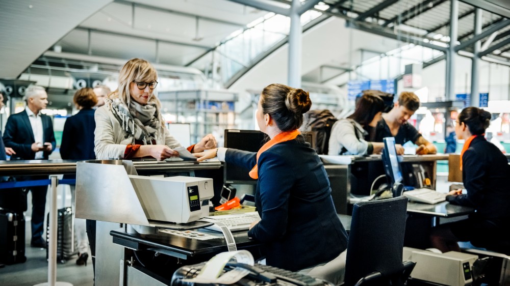 A female international student in a scarf and thick-rimmed glasses, handing her ticket over to airport staff