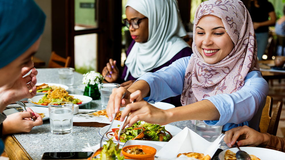 Three female international students wearing hijab sit at a table with salads and halal food at their US university dining hall.