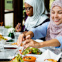 Three female international students wearing hijab sit at a table with salads and halal food at their US university dining hall.