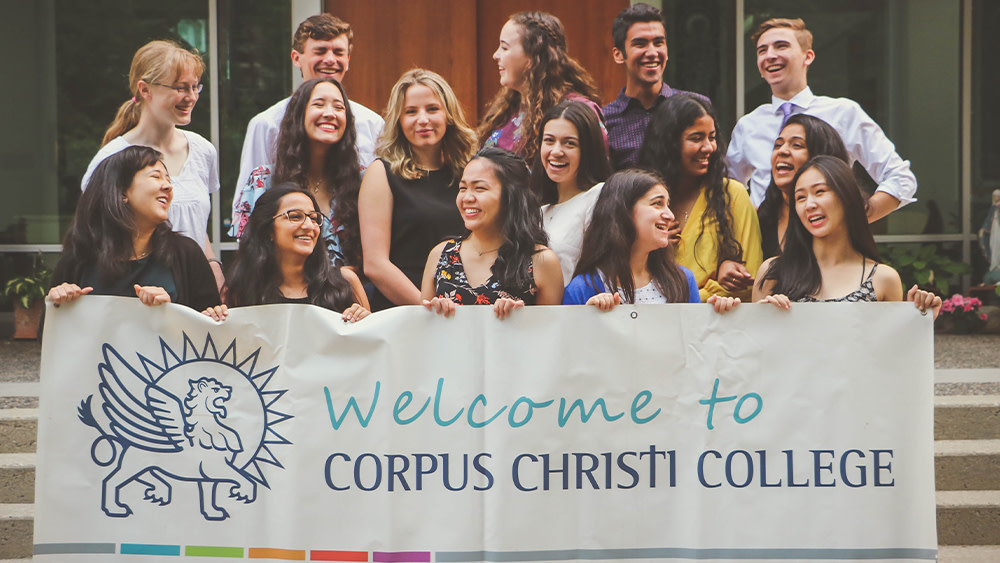 A group of international students stand on stairs on the UBC campus in Vancouver and hold up a banner saying Welcome to Corpus Christi College