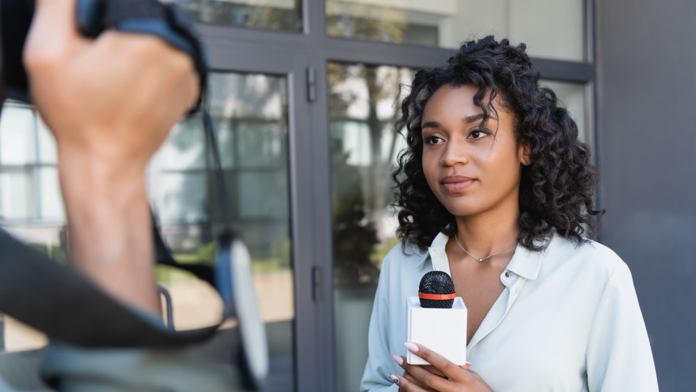 A female international student taking journalism courses at a Shorelight US university holds a microphone and reports a breaking news story for a news camera.