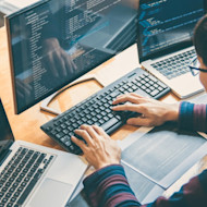 View from behind of a young man working at a desktop monitor and laptop, with both displaying computer code.