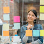A female international student entrepreneur major stands in front of a glass wall covered in colorful sticky notes at her internship, working on answering what is a entrepreneur and listing the skills of entrepreneurs for her upcoming project.