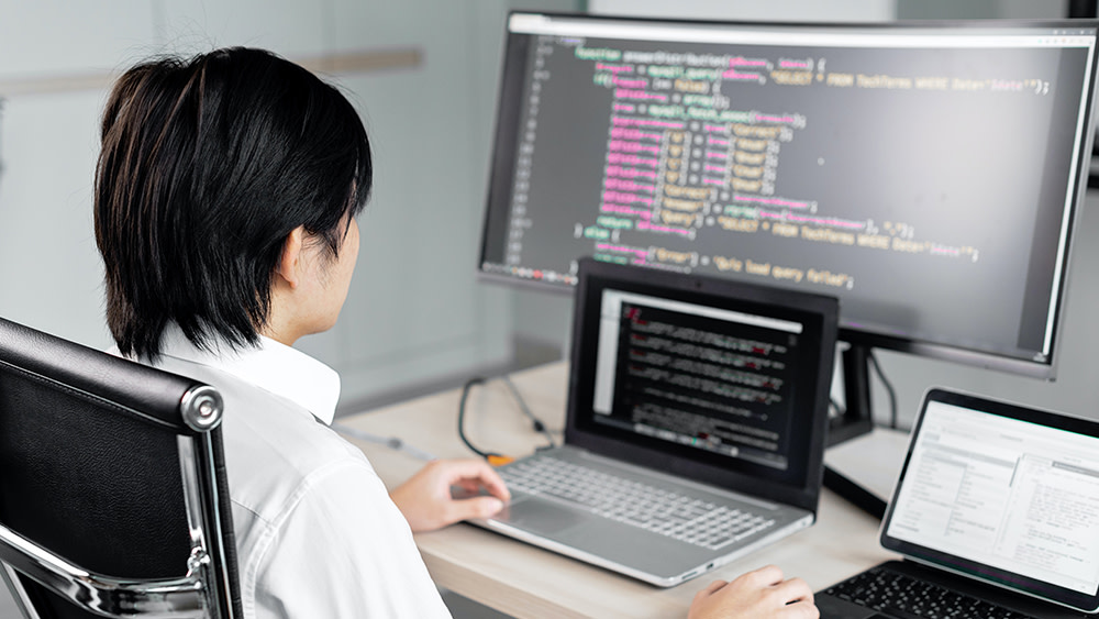 An Asian international student studying cyber security sits in front of a laptop and second monitor and studies code for cyber security types, security breaches, and how to define cyber security