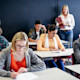 This photo shows university students taking the GMAT exam, with college students writing in GMAT test booklets with a female professor standing behind them.