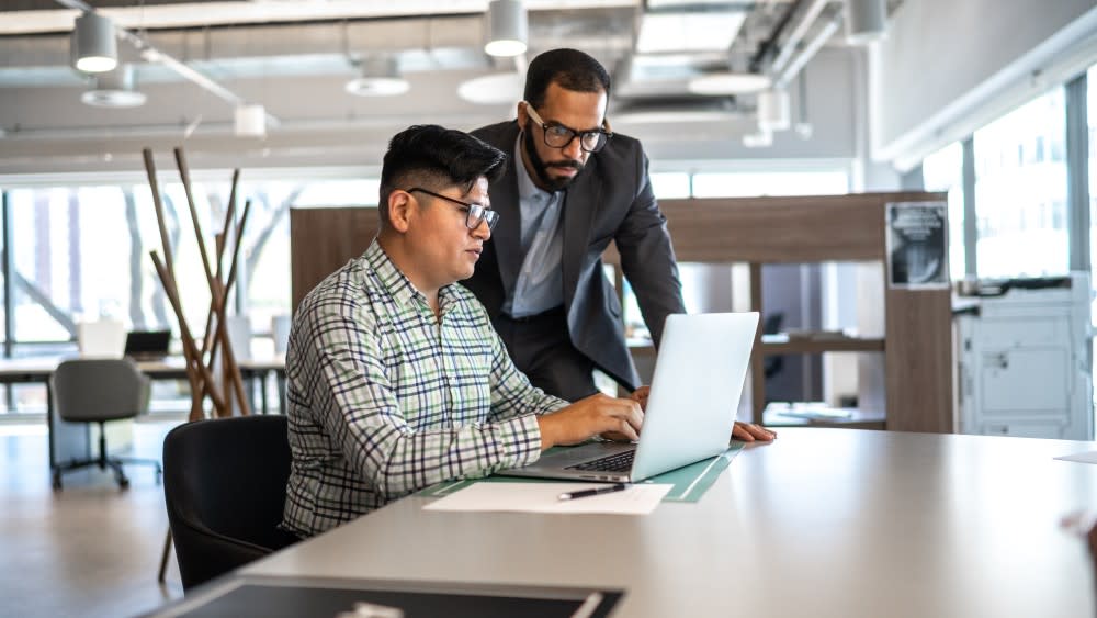 A male international student from South Asia sits in front of a laptop while his mentor, a man in a suit jacket and glasses, looks over his shoulder