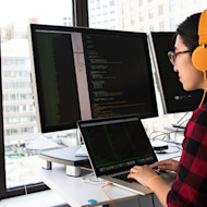 A female international student from Asia wearing headphones sits at her internship desk in front of windows showing a city skyline and codes on a laptop in front of two external monitors
