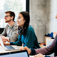 A male and a female international student sit in front of laptop computers at a long table in a US university classroom, focusing on curriculum covering what are technical skills, technology skills for students, and technical skills to learn.
