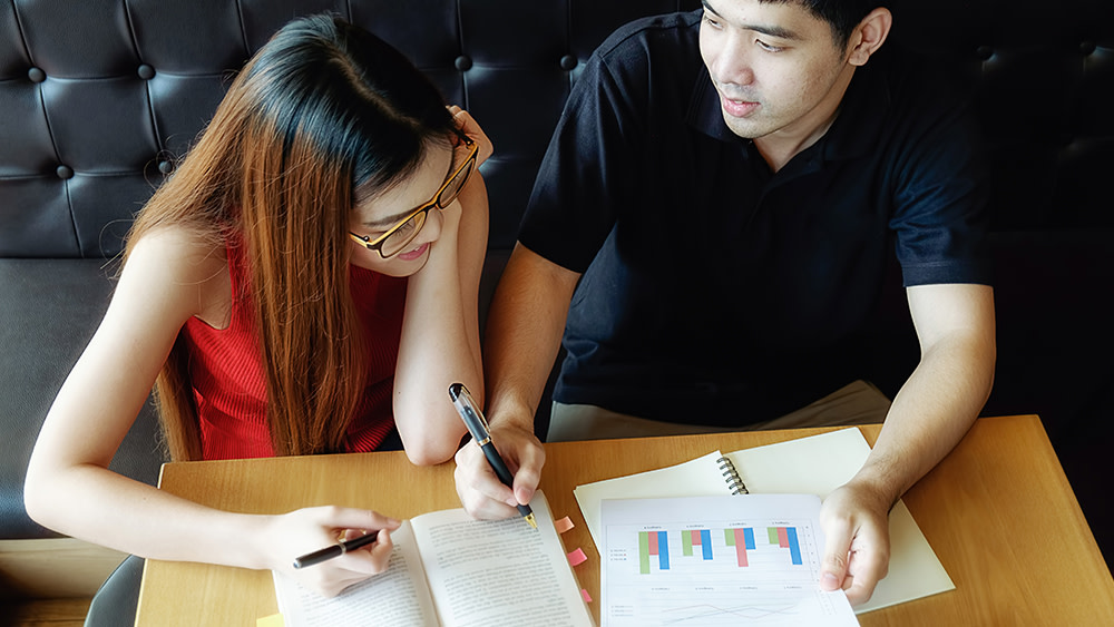 Two international students studying statistics sit at a table with a notebook and chart printout to complete homework for their statistics courses.