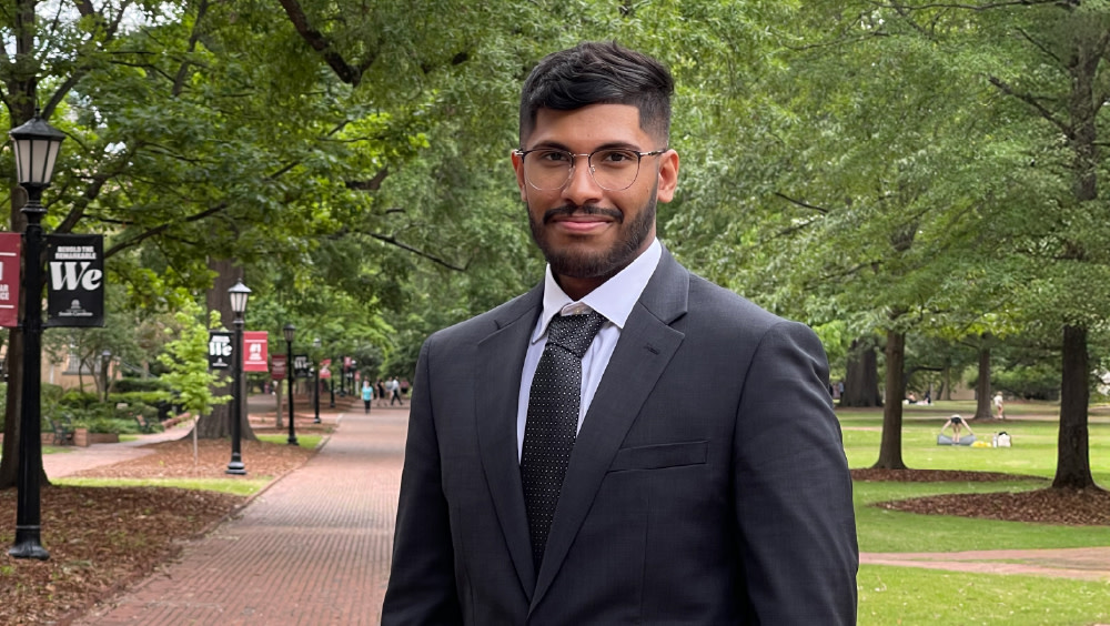 Mustafa, an international student from Saudi Arabia, stands in a suit and tie on the University of South Carolina campus and smiles for the camera.
