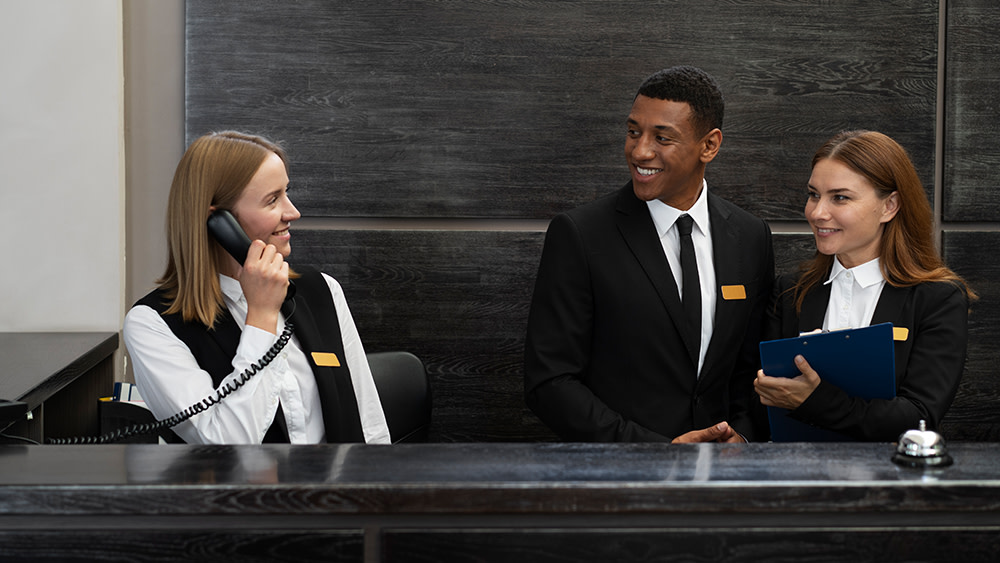 A hotel manager and two hotel management student intern employees work at the reception desk of a hotel.