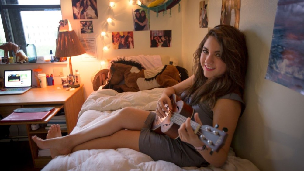 A female international student sits on her dormitory bed and plays acoustic guitar in her student accommodations at her US university.