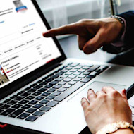 This photo shows an open laptop with a "us university rankings" article showing on the screen and a hand on the mousepad and a different hand pointing to the screen. The users are researching US top universities and US university ranking listings.