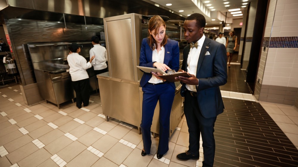 Two international students in uniform suits stand in a commercial kitchen and compare notes during their hospitality internship