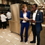Two international students in uniform suits stand in a commercial kitchen and compare notes during their hospitality internship