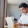 A male international student sits in front of a laptop for his online classes and online college courses at his US university