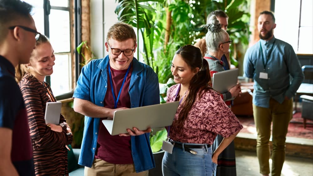 A female international student participating in a Shorelight Career Accelerator internship stands next to her male colleague holding a laptop in front of potted trees in an office
