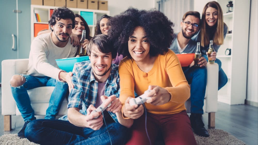 Facing the screen, a man and woman are playing a video game while a group of friends watches behind them.