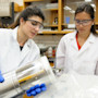 Two female international students wearing lab coats and goggles work on research at a lab at UMass Boston.