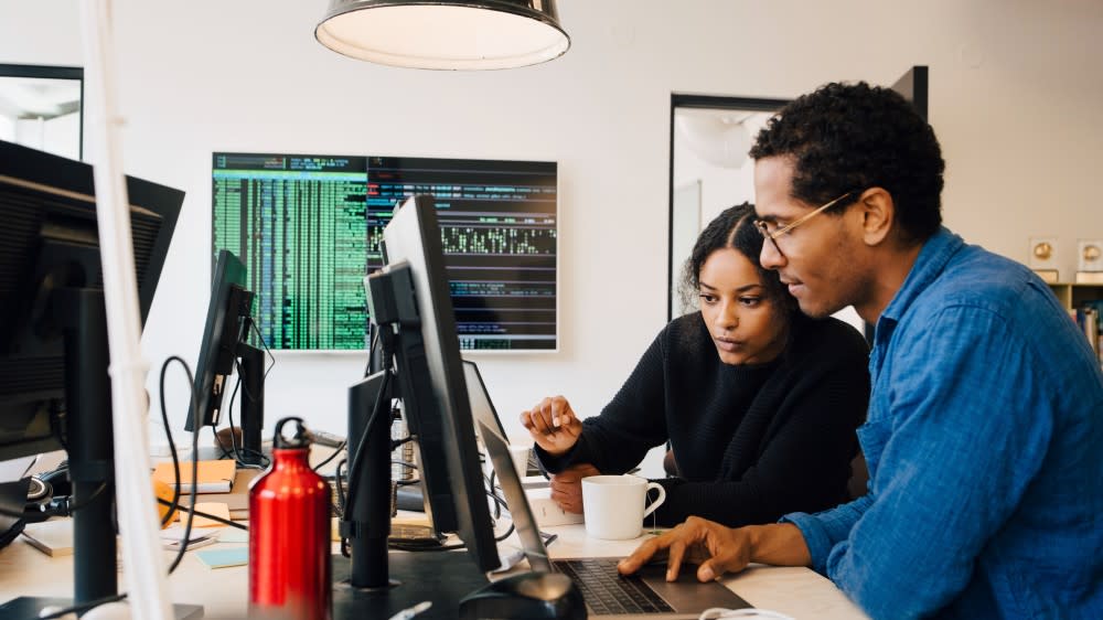 Two international students sit side by side at a table in front of a computer monitor near a blackboard