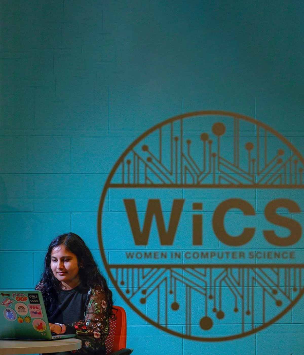 A woman sitting working on a laptop with a "Women in Computer Science" mural behind her.