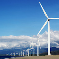 Wind turbines in a row line up before a mountain range and blue sky with white clouds