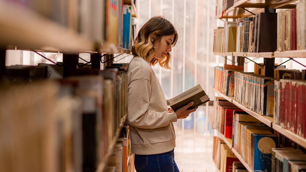 A female international student majoring in English literature stands among the stacks in her US university library and reads an open book.