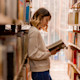 A female international student majoring in English literature stands among the stacks in her US university library and reads an open book.