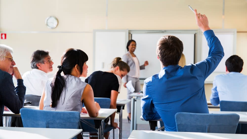 A man in a classroom sitting with his back to the camera, raises his hand, with the instructor facing the class up front.