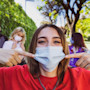 A female international student stands outside on a US university campus on a sunny day and points to the face mask she's wearing while two fellow masked students stand behind her