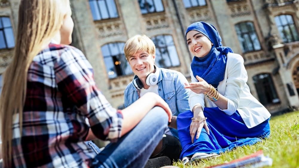 A blond male international student with headphones and a female international student wearing a blue burka sit outside across from a male student sitting cross-legged on the lawn of a college campus quad.
