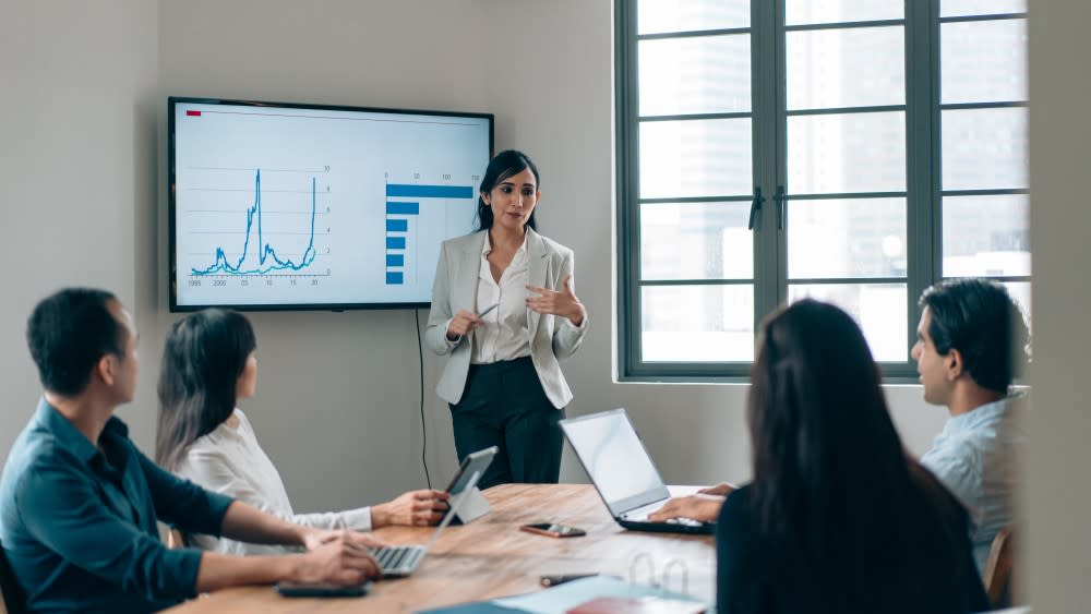 A female international student studying for her master of business analytics with UDayton Global stands in front of a business presentation in a conference room in front of a table of colleagues