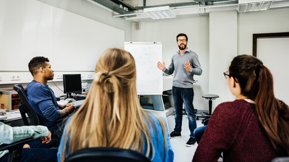 A professor stands next to an easel in front of two international students seated in a US university classroom. The students are learning what is a minor in planning their majors and minors for their degree programs.