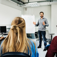 A professor stands next to an easel in front of two international students seated in a US university classroom. The students are learning what is a minor in planning their majors and minors for their degree programs.